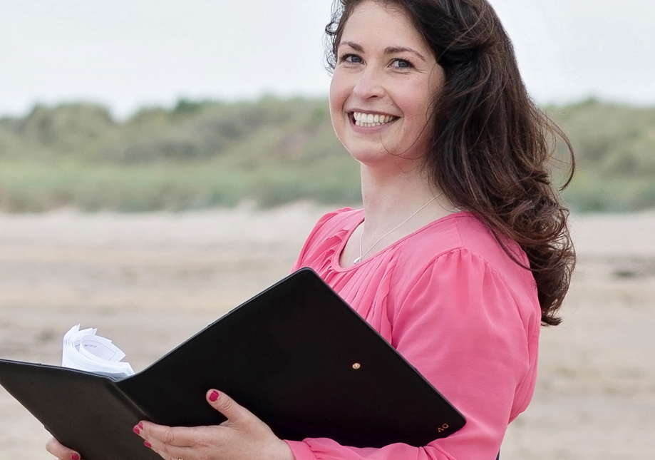 A woman with long brown hair is staring into the camera with a smile. She is dressed in pink and holding a black folder. She is standing on a beach.